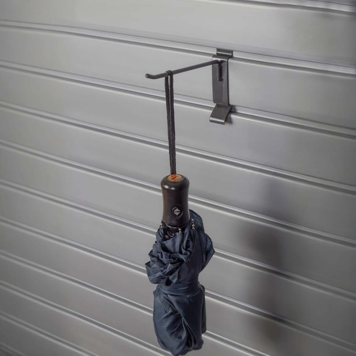 Slatwall Hooks holding a folded black umbrella by its strap on a ribbed gray slatwall panel