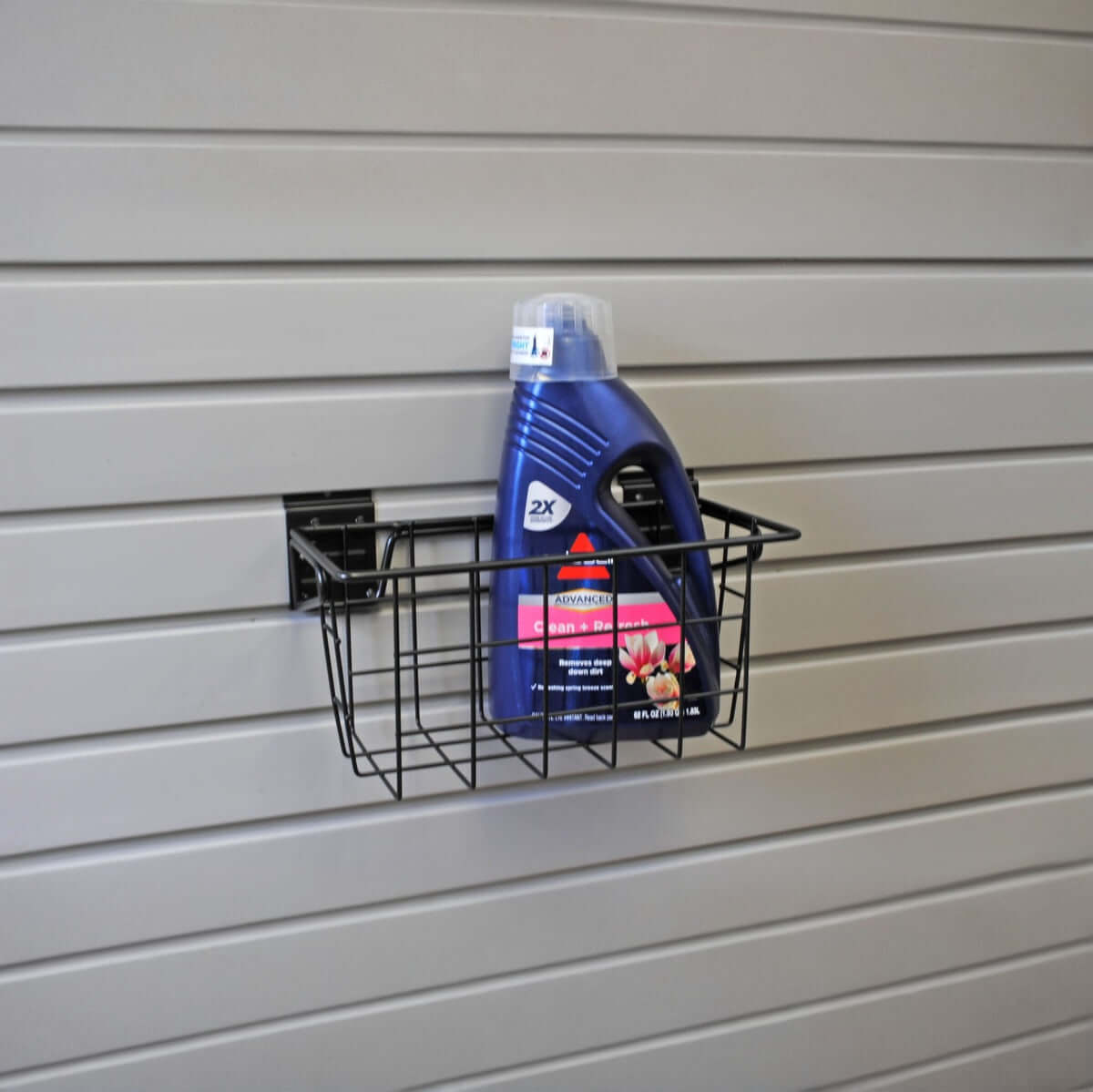 Slatwall Baskets wire narrow basket mounted on gray slatwall panel holding a large blue Bissell laundry detergent bottle